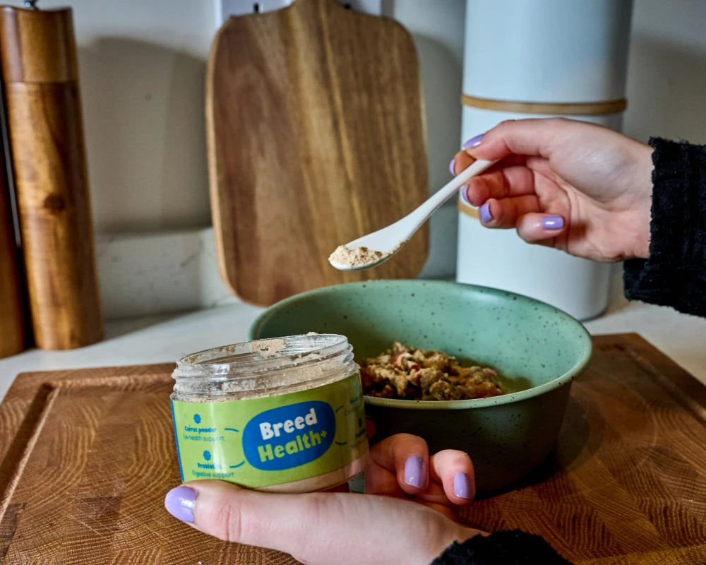 Person holding a jar of 'Bread Health+' and a bowl with a spoonful of food on a kitchen counter.