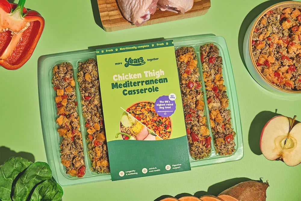 A vibrant magenta background with a round bowl on the left filled with a colorful mix of diced steak, kidney, mushrooms, quinoa, red pepper, carrot, potato and greens. On the right is a green three-compartment meal tray partially covered by a matching magenta sleeve labeled “Years – Steak & Kidney Mushroom Hotpot, personalised for Hugo,” showing the same chunky hotpot portions.